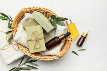 Handmade olive soap bars, essential oil bottles, and fresh olive branches arranged in a wicker basket on a white tile background, evoking a tranquil spa settingの写真素材