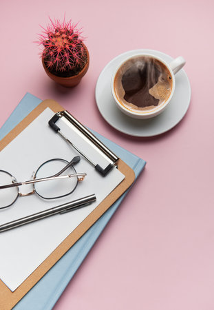 Clipboard, eyeglasses, pen, coffee cup and cactus plant are lying on pink surface, composing a relaxing break time sceneの写真素材