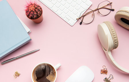 Flat lay of a pastel pink desk with coffee, headphones, keyboard, mouse, glasses, notebook, pen,...