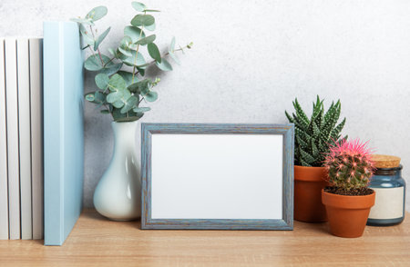 Empty blue frame mockup standing on wooden desk with eucalyptus, succulents, books and candle on...