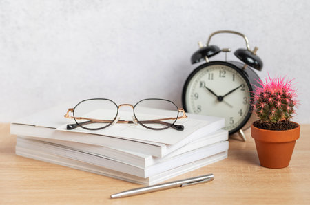 Eyeglasses sit atop a stack of notebooks beside a pen, alarm clock, and small cactus plant, creating a serene workspace ambianceの写真素材