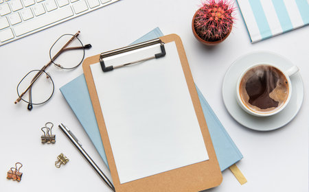 Home office desk workspace with blank clipboard, keyboard, coffee, cactus, glasses, pen, notebook, and binder clips, inviting creativity and productivityの写真素材