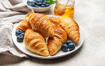 Delicious golden croissants are served on a white plate with fresh blueberries and honey for a sweet breakfastの写真素材