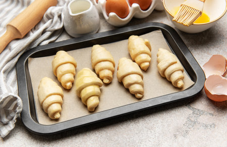 Raw croissants on baking sheet, ready for the oven, with ingredients and tools around