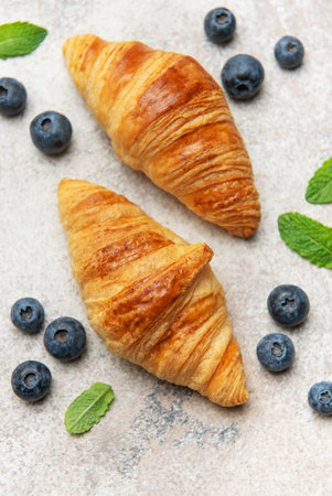 Two golden brown croissants are laying on a textured surface next to fresh blueberries and mint leaves, creating a tempting breakfast sceneの写真素材