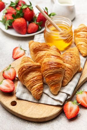 Freshly baked croissants are served on a wooden board with strawberries and honey for a delightful breakfastの写真素材