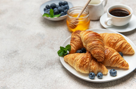 Freshly baked croissants are served on a white plate with blueberries, honey, and a cup of coffee, creating a tempting breakfast sceneの写真素材