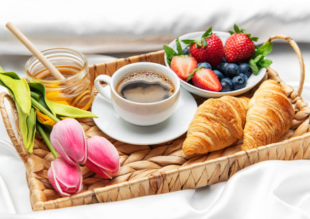 Delicious breakfast tray with coffee cup, croissants, fresh berries, honey and pink tulips, served in bed on white sheetsの写真素材