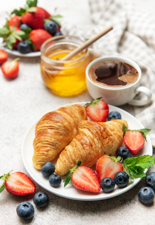 Freshly baked croissants are served on a plate with juicy strawberries, blueberries, mint leaves, a cup of coffee and honey for a delightful breakfastの写真素材