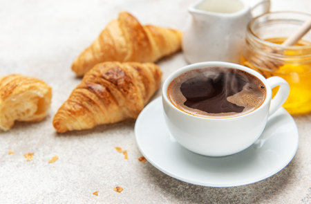 Freshly baked croissants, steaming coffee, honey, and milk on a table create a tempting breakfast sceneの写真素材