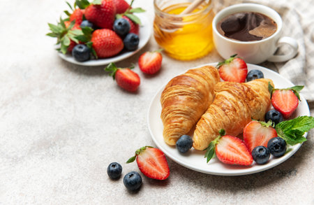 Freshly baked croissants are served on a plate with juicy strawberries, blueberries, mint leaves, a cup of coffee and honey for a delightful breakfastの写真素材