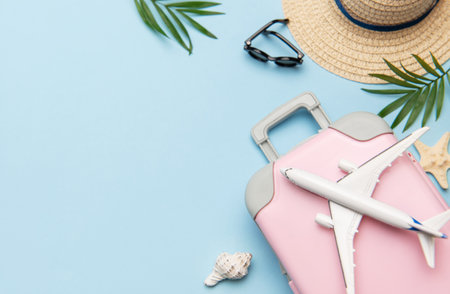 Summer travel planning concept with pink suitcase, toy airplane, sunglasses, straw hat, seashells and palm leaves on blue background with copy spaceの写真素材