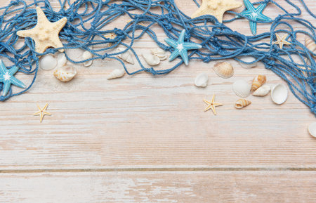 Blue fishing net, seashells, and starfishes arranged on light wooden planks create a charming seaside borderの写真素材
