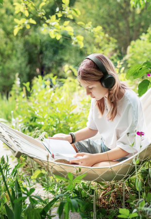 Teenager girl relaxing in a hammock in the garden, reading a book and listening to music with headphonesの写真素材