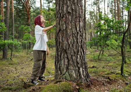 Young woman with pink hair stands in a lush forest, gently touching a large tree trunk, appreciating the peace and beauty of natureの写真素材