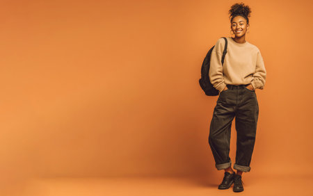 Studio portrait of a smiling young student wearing casual clothes, posing with a backpack on an orange backgroundの素材