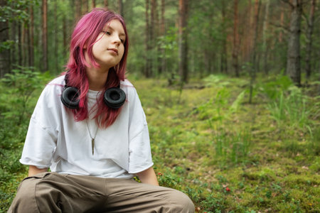 Teenager with pink hair enjoying tranquility in a forest setting, headphones resting on shoulders