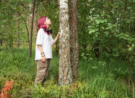 Teenager with headphones enjoying nature in a green forest, touching a birch tree trunk