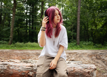 Teenager with pink hair enjoying music in nature, sitting on a tree trunkの写真素材