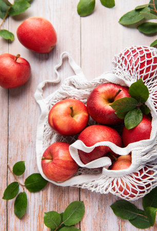 Ripe red apples with leaves are inside a reusable shopping bag on a wooden table, symbolizing a sustainable lifestyleの写真素材