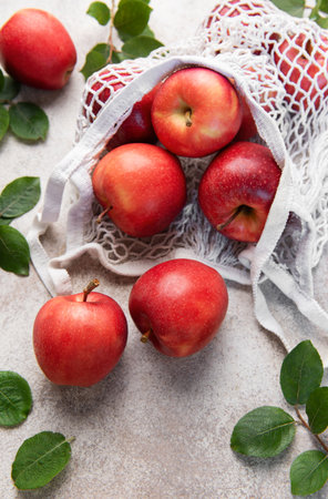 Ripe red apples are spilling out of a reusable white mesh bag onto a textured surface, promoting healthy eating and sustainabilityの写真素材