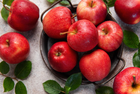 Vibrant red apples with water droplets, nestled amongst green leaves in a rustic metal basket,...