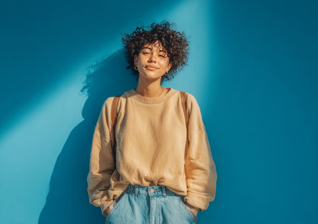 Confident young woman with curly hair standing against a blue wall in bright sunlightの素材