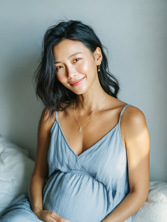 Expectant asian woman gently touching her baby bump, smiling with serene expressionの素材