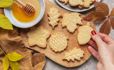 Hand holding a leaf shaped cookie among other autumn inspired shortbread and a bowl of honeyの写真素材