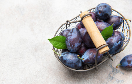 Ripe organic plums with green leaves in a rustic basket and scattered on a gray surfaceの写真素材