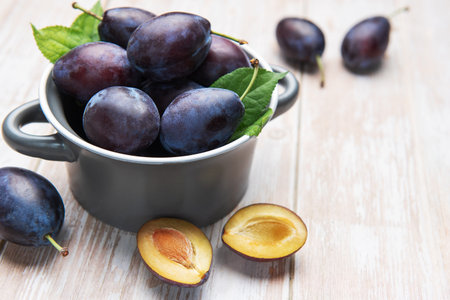 Fresh ripe plums and green leaves filling a grey bowl on a light wooden tableの写真素材