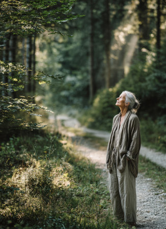 Senior woman standing on a forest path enjoying the sunlight and serene natureの素材