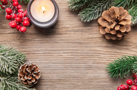 Illuminating candle with pine cones, berries, and frosted fir branches on wooden surface