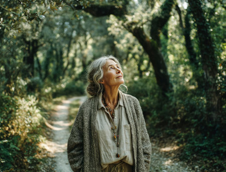 Senior woman standing on a path in a forest looking up at the tree canopyの素材