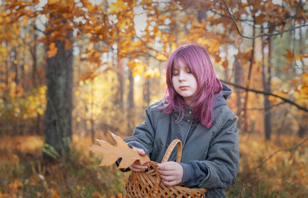 Young girl with dyed pink hair holding a yellow oak leaf and basket in an autumn forestの写真素材