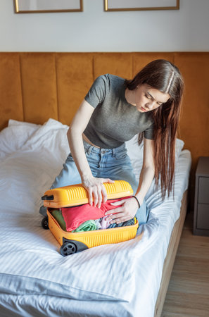 Young woman packing an overflowing yellow suitcase for a trip while sitting on a bedの写真素材