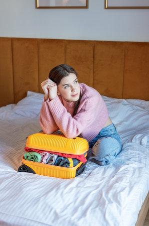 Young woman packing suitcase on bed, looking up and contemplating travelの写真素材