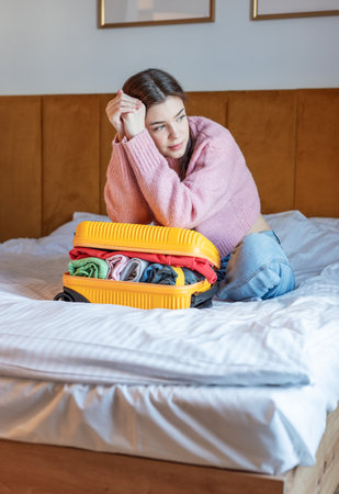Woman feeling thoughtful while packing clothes into a yellow luggage on a bedの写真素材