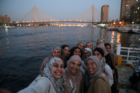 Group of Egyptian women enjoying selfies by the Nile river during twilight, highlighting their joyful expressions and the beautiful urban backdrop of the cityの素材