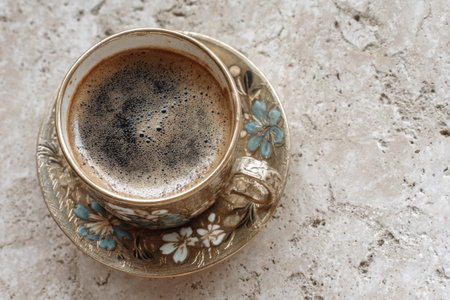 Ornate Turkish coffee cup filled with dark brew sits on a decorative saucer, highlighting intricate floral patterns against a neutral textured backgroundの素材