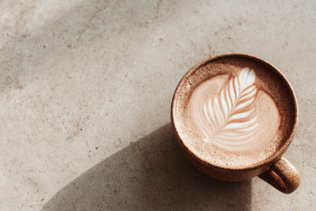 A beautifully crafted latte with leaf art in a brown ceramic cup, resting on a textured surface, creating a serene coffee moment with copy spaceの素材