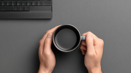 Hands of a businessman grasping a black ceramic high fidelity mug on a gray desk, accompanied by a modern keyboard, creating a focused work environmentの素材