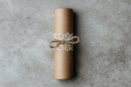 Cylindrical kraft paper gift tied with lace and twine, resting on a gray textured background, highlighting minimalist design and charming aestheticsの素材