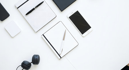Flat lay of essential modern office supplies including a white notebook, black pen, smartphone, and over-ear headphone arranged neatly on a bright white surface for a clean, minimalist look.の素材