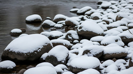 Snow covered river rocks line the bank of a calm dark water stream in a winter landscapeの素材