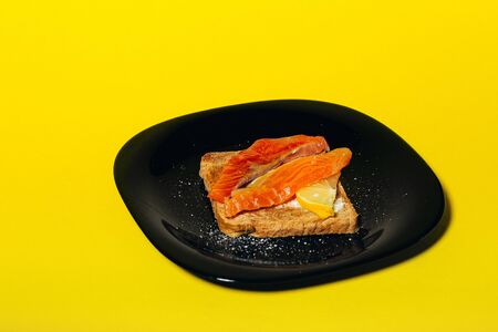 tasty healthy toast with butter, salmon and lemon on a black glossy plate on a yellow background. top side viewの写真素材