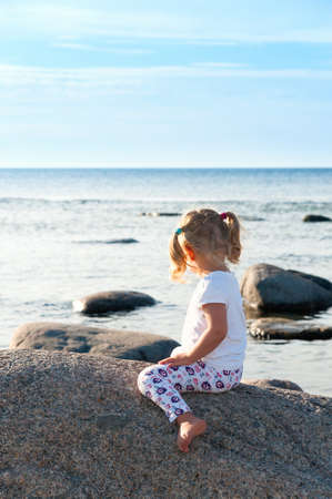 Rear view of a little girl straddling a boulder on the beach as she sits in the summer sunshine looking out to seaの写真素材