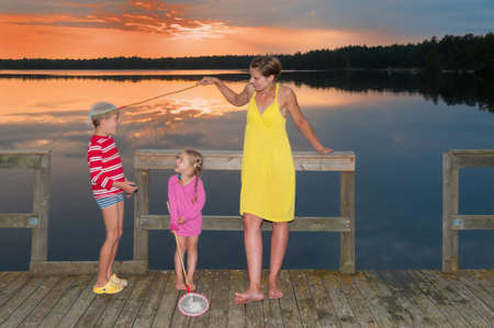 Young mother standing with her children on a wooden pier at a lake in Smaland, Sweden, in the light of an amazing sunset and kidding with her little son and catch him with a landing net  の写真素材