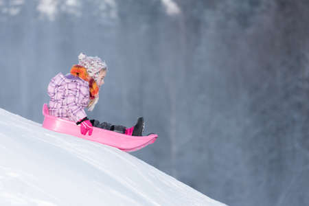 Little girl sledging downhill on having fun. Pink sledge and jacket, colorful scarf.の写真素材