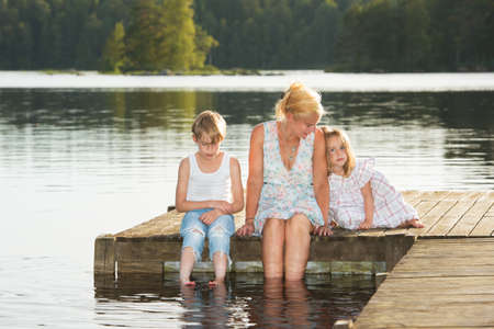 Beautiful young mother and her two kids sitting on a jetty in the summer sun, bathe the feet in the water, and enjoy a beautiful day in midsummer.の写真素材
